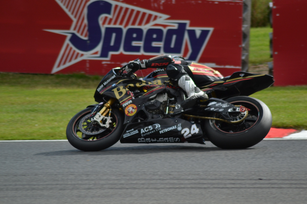 Eine Person in Helm und Schutzkleidung fährt ein Motorrad auf einer Rennstrecke, umgeben von Gras und einem Schild im Hintergrund.