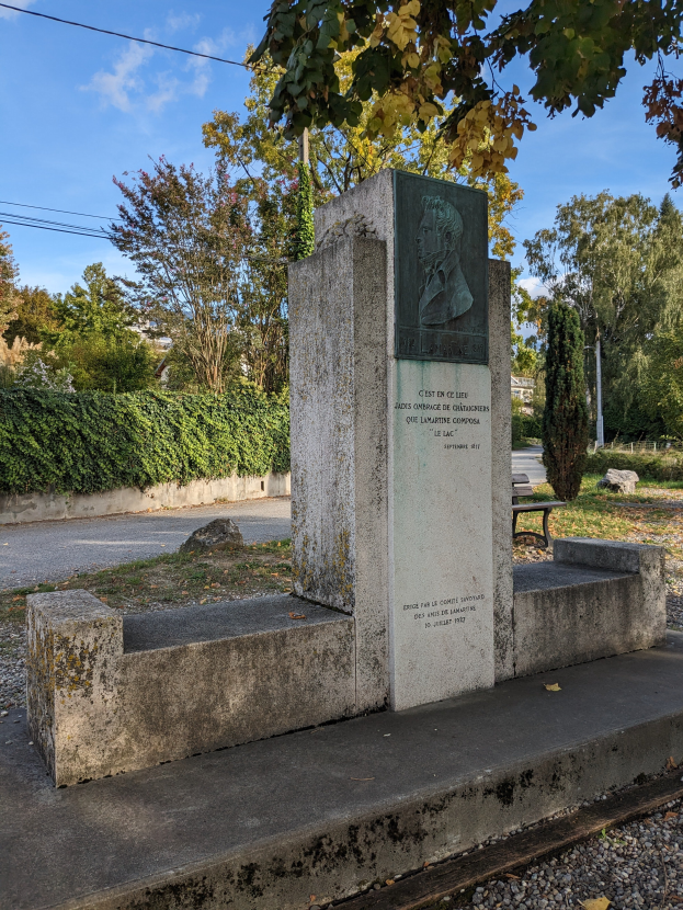 Ein steinernes Denkmal mit eingraviertem Text steht in einem Park, umgeben von Gras, Pflanzen, Bäumen und einer Bank davor, mit einer bewölkten Himmel.