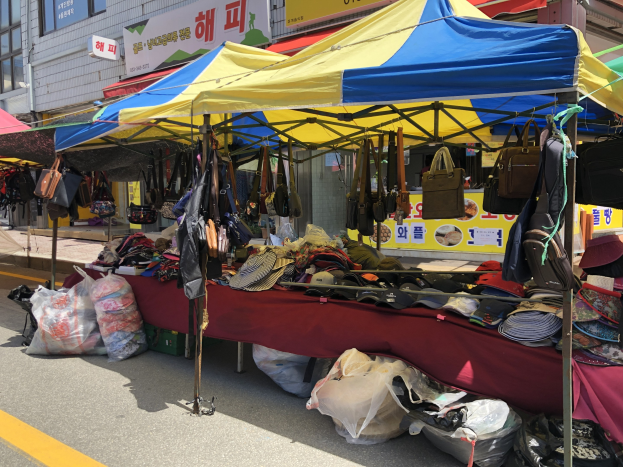 Ein Straßenmarkt mit Zelten, die verschiedene Gegenstände wie Taschen und Hüte präsentieren, vor Häusern mit Fenstern und Texttafeln im Hintergrund.