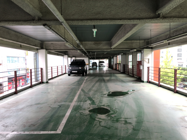 Ein mehrgeschossiges Parkhaus mit parkenden Autos, Gel├Ąndern, S├Ąulen, Lichtern, Rohren und B├Ąumen, vor einer Stadtkulisse mit Fenstern.