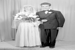 Ein Mann und eine Frau stehen Seite an Seite auf einer Bühne, lächeln und halten ein Blumenbouquet, mit einem Vorhang dahinter.