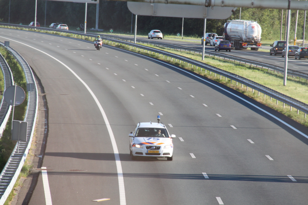 Ein Auto auf einer Autobahn mit einem stationären Polizeiwagen am Rande, andere Fahrzeuge und Bäume, Gelönder, Pfosten und Planken im Hintergrund.