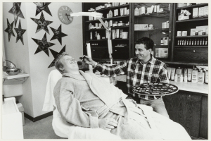 Ein Mann lächelt in einem Zahnarztstuhl, während ein anderer Mann neben ihm steht und eine Platte hält, mit einem Waschbecken, Sternen, einer Uhr und Regalen voller Flaschen im Hintergrund.