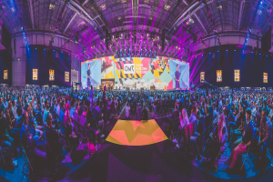 Eine große Menschenmenge sitzt auf Stühlen vor einer hell erleuchteten Bühne mit mehreren stehenden Individuen, wahrscheinlich bei einem Online-Marketing-Event.