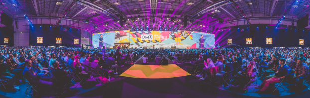 Eine große Menschenmenge sitzt auf Stühlen vor einer hell erleuchteten Bühne mit mehreren stehenden Individuen, wahrscheinlich bei einem Online-Marketing-Event.