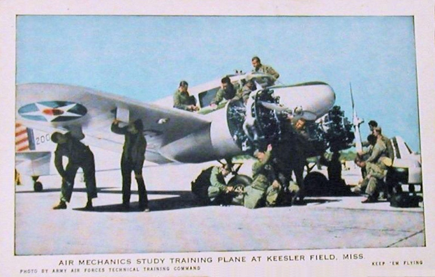 Eine Gruppe von Menschen um ein Flugzeug auf einer Landebahn herumstehend mit einem klaren blauen Himmel im Hintergrund und Text unten lesend "Air Mechanics Study Training Plane at Keesler Field, Miss."