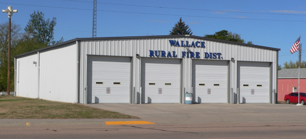 Feuerwache des Wallace Rural Fire District mit einer Straße darunter, Gras links, einem Lichtmast rechts, Bäumen im Hintergrund, einem zentralen Turm und einer Flagge, die darüber weht, unter einem sichtbaren Himmel.