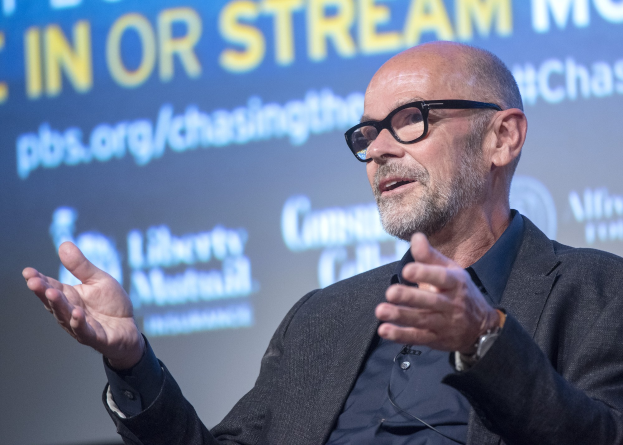 Steve Ballmer, Gründer und CEO von PBS, spricht in einen Mikrofon in einem Anzug und Brille, mit einem Bildschirm, der Text anzeigt, im Hintergrund.