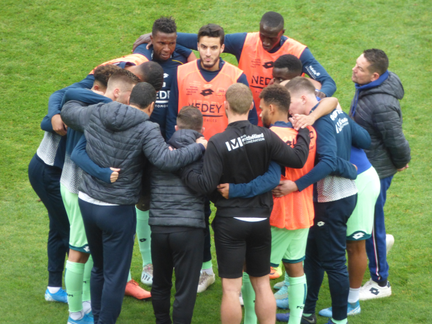Eine Gruppe von Männern in Jacken steht zusammen im Kreis auf einem Fußballfeld.