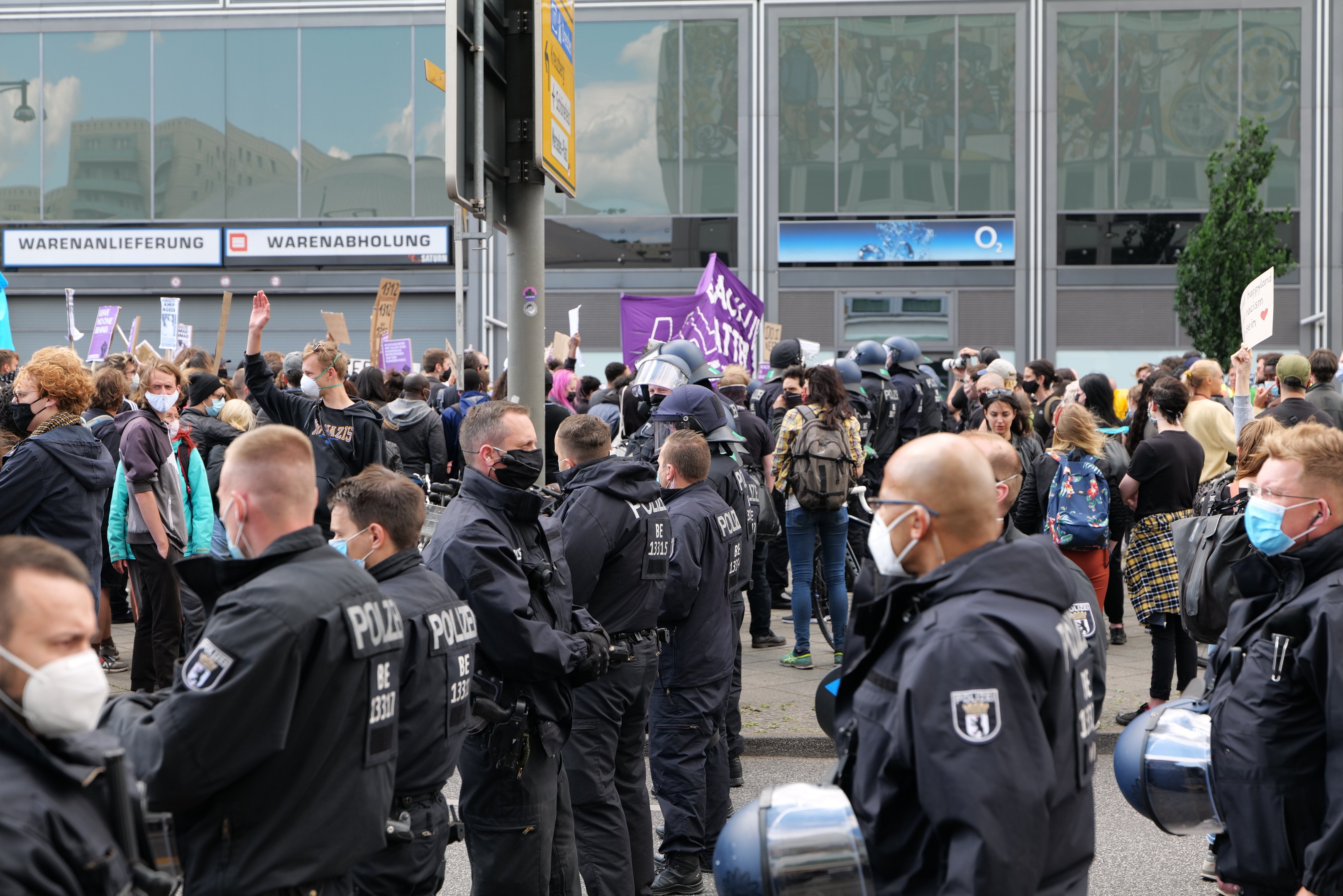 Eine große Gruppe von Menschen steht vor einem Gebäude und hält Schilder hoch, trägt Helme und hat eine Stange und ein Schild im Vordergrund und einen Baum im Hintergrund, was auf eine Demonstration hindeutet.