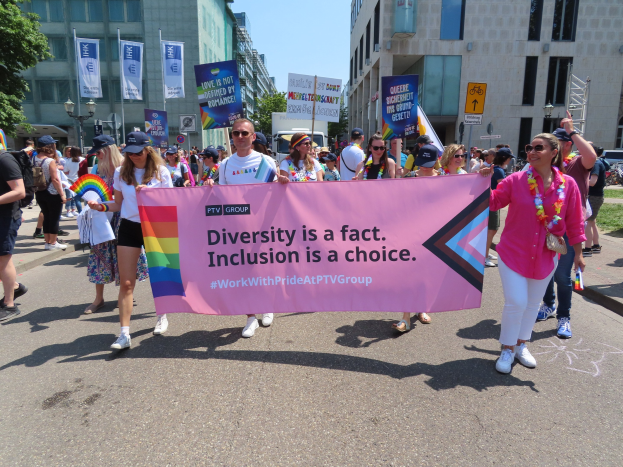 Eine Gruppe von Menschen marschiert auf einer Straße, hält ein Banner mit der Aufschrift "Diversität ist eine Tatsache, Inklusion ist eine Wahl" und trägt teilweise Mützen und Schutzbrillen, während andere zusätzliche Banner halten, vor einem Hintergrund aus Gebäuden, Bäumen und einem klaren Himmel.