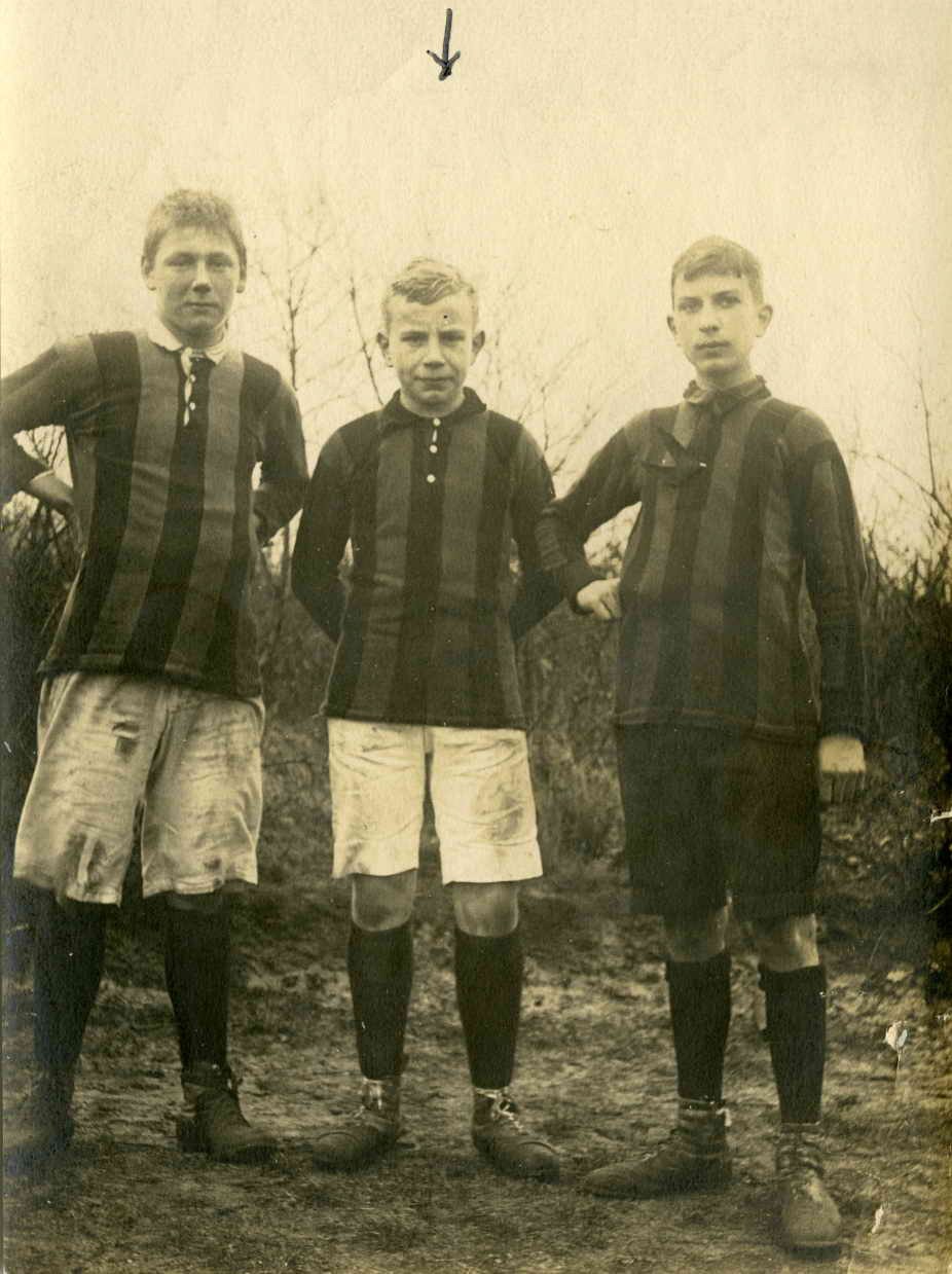 Drei junge Männer in Fußballtrikots stehen Seite an Seite auf einem Feld mit Bäumen und einem klaren Himmel im Hintergrund.