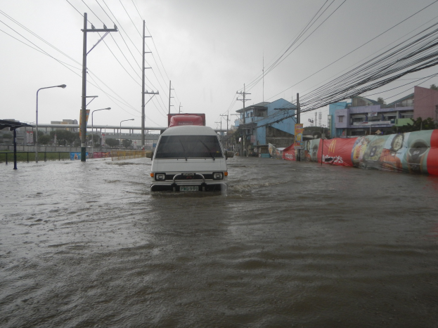 Ein Van fährt durch eine überflutete Stadtstraße mit Strommasten, Laternenmasten und Gebäuden auf beiden Seiten, eine Brücke im Hintergrund und der Himmel oben im Bild.