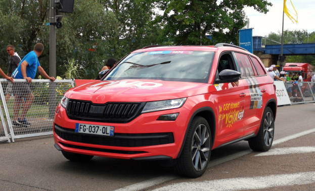 Ein rotes Auto fährt auf einer Straße neben einer Menschenmenge, einige tragen Mützen, mit Maschendraht auf der linken Seite, einem Verkehrssignalmast, Bäumen, einer Brücke, einem Fahnenmast und einem bewölkten Himmel im Hintergrund.