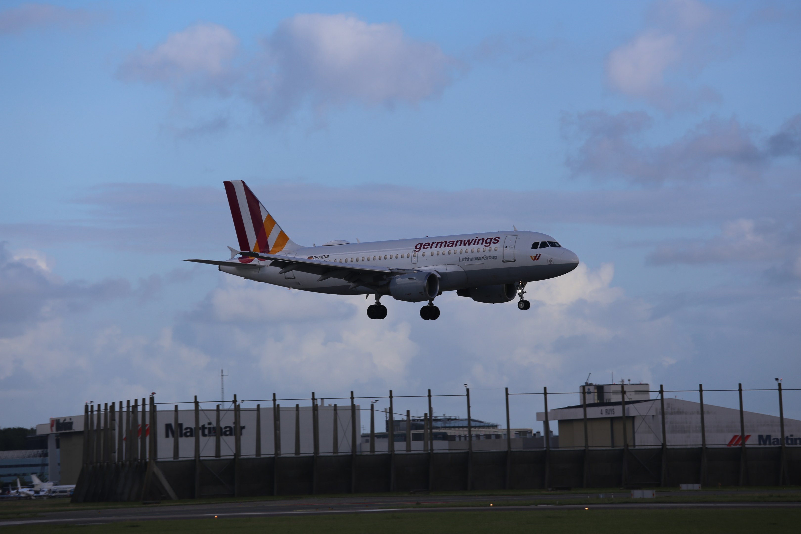 Germanwings Airbus A320-200 beim Start am Frankfurter Flughafen, mit Gras darunter, einem Zaun im Vordergrund, Gebäuden und Bäumen im Hintergrund und einer bewölkten Himmel.