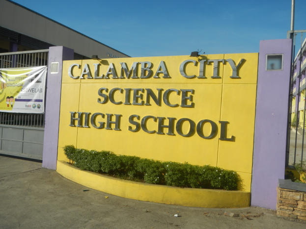 Gebäude der Calamba City Science High School mit einem Namensschild, Pflanzen, einer Fahne, einem Tor, einem Pfahl und einem klaren blauen Himmel im Hintergrund.