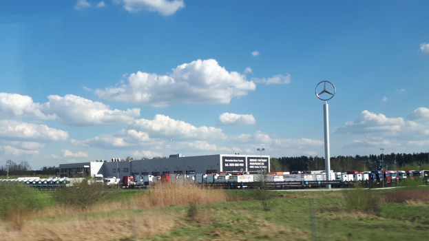 Außenansicht einer Mercedes-Benz-Fabrik in Deutschland mit Fahrzeugen auf der Straße, Grünfläche, Gebäuden und einem Logo-Pfahl unter einem bewölkten Himmel.