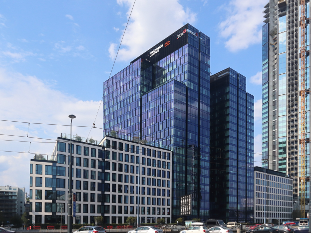 Großes Gebäude mit vielen Fenstern, als Deutsche Bank Hauptsitz in Frankfurt, Deutschland identifiziert, umgeben von städtischer Infrastruktur einschließlich Straßenlaternen, Strommasten, Stromkabeln, Kraftfahrzeugen, Grünflächen und einem bewölkten Himmel.