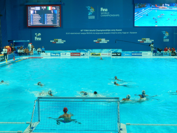 Wasserballspieler bei der 2016 FINA Weltmeisterschaft in Kazan im Pool gegeneinander antretend, mit Zuschauern auf der linken Seite und einem Scoreboard im Hintergrund.