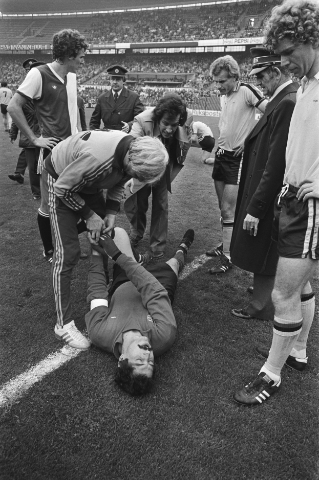 Schwarzes und weißes Bild eines Fußballspielers, der auf dem Boden liegt und von einer Gruppe Männer umgeben ist, mit einer Stadionkulisse im Hintergrund.