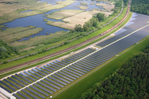 Eine Luftaufnahme eines Solarparks mit zahlreichen Solarmodulen auf einem Feld, umgeben von Bäumen, Gras und Wasser, mit einem Zug, der auf einem nahen Bahnsteig fährt.