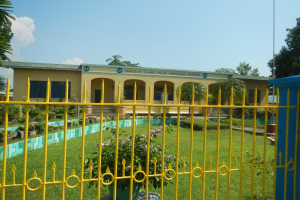 Ein gelbes Schulgebäude mit einem passenden Zaun, umgeben von grünem Gras, Pflanzen und Bäumen, unter einem Himmel voller weißer, flauschiger Wolken.