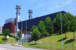 Großes Industriegebäude mit vielen Fenstern, als Kraftwerk identifiziert, umgeben von Straßeninfrastruktur, Fahrzeugen, Grünflächen und einem bewölkten Himmel.