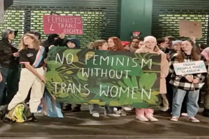 Eine Gruppe von Menschen mit einem Banner, auf dem "Kein Feminismus ohne Trans-Frauen" steht und Schilder in einer öffentlichen Fläche mit einer Wand und einem Rollladen im Hintergrund.