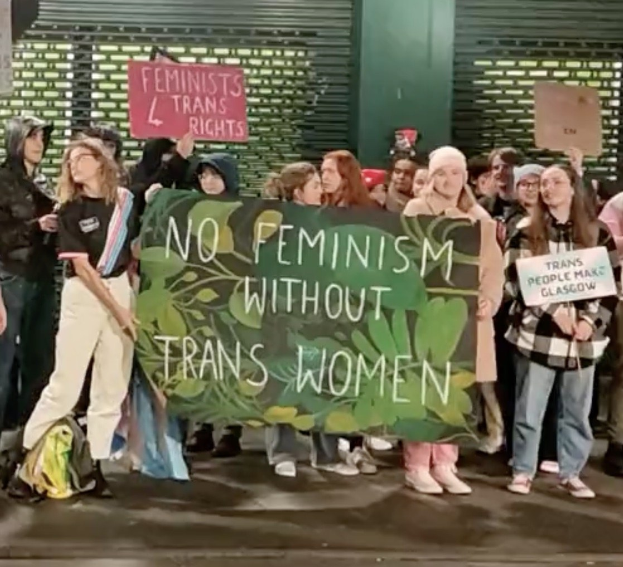 Eine Gruppe von Menschen mit einem Banner, auf dem "Kein Feminismus ohne Trans-Frauen" steht und Schilder in einer öffentlichen Fläche mit einer Wand und einem Rollladen im Hintergrund.
