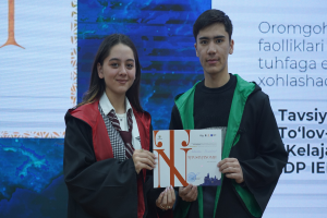 Ein Mann und eine Frau in Graduationsroben stehen nebeneinander, jeder hält ein Zertifikat in der Hand, mit einem Bildschirm mit Text dahinter.