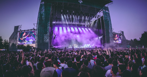 Eine große Menschenmenge versammelte sich auf einem Musikfestival, mit einer Bühne in der Mitte, auf der Musiker Instrumente spielen, beleuchtet von Scheinwerfern und umgeben von Bildschirmen, mit einem Gebäude, Bäumen und einem klaren blauen Himmel im Hintergrund.