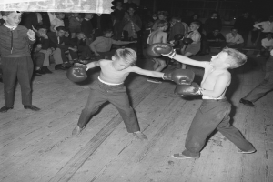 Zwei junge Jungs in Boxhandschuhen kämpfen in einem Boxkampf, umgeben von einer Gruppe von Menschen, die im Hintergrund stehen und sitzen, dargestellt in einem Schwarz-Weiß-Foto.