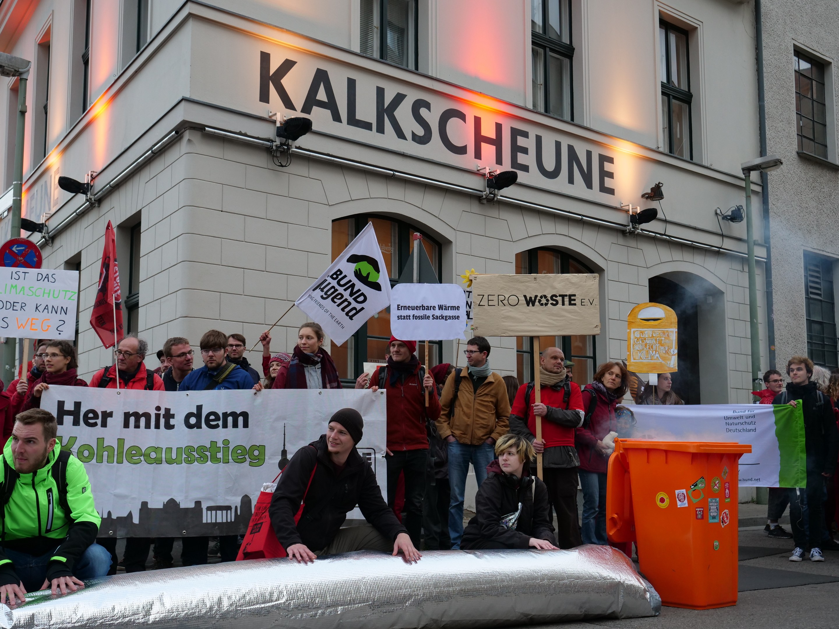Eine Gruppe von Menschen mit Protestschildern vor einem Gebäude mit einem Müllcontainer auf der rechten Seite und sitzenden Personen im Vordergrund, was auf eine Protestaktion in Deutschland hinweist.