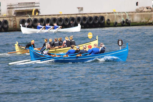 Eine Gruppe von Menschen in einem blauen und gelben Boot auf dem Wasser, die Paddel halten, mit einer Wand mit Reifen und einem Gebäude im Hintergrund, was auf eine Regatta hindeutet.