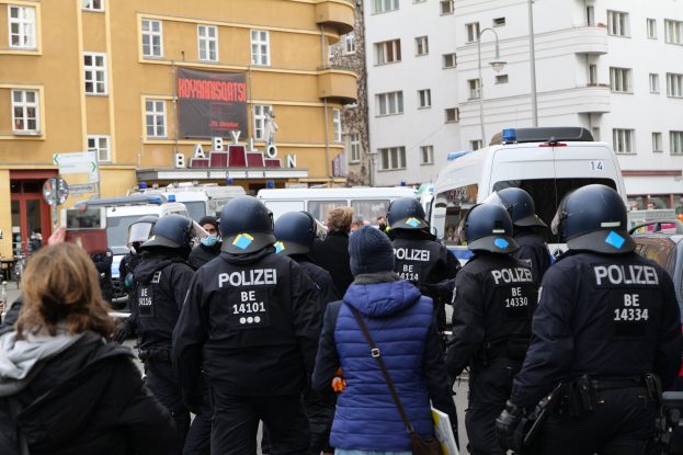 Eine Gruppe von Polizisten steht vor einer Menge von Menschen, die Helme und Jacken tragen, während einer Demonstration in Berlin, Deutschland, mit Fahrzeugen, Gebäuden, Laternenpfählen und einem Banner mit Text im Hintergrund und einer Person mit einer Kamera.