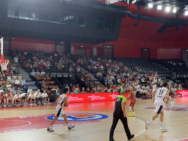 Eine Gruppe von Menschen, die Basketball in einer Turnhalle spielen, mit einem Basketballkorb auf der linken Seite und Zuschauern im Hintergrund, unter hellen Stadionlichtern.