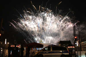 Ein Polizeiwagen steht vor einer Menschenmenge, im Hintergrund sind Gebäude und Bäume beleuchtet, während farbige Feuerwerke den Himmel erhellen, während des Silvesterfeuerwerks in Berlin.