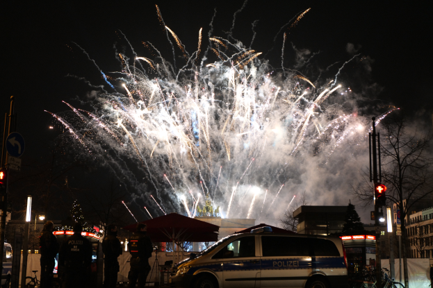 Ein Polizeiwagen steht vor einer Menschenmenge, im Hintergrund sind Gebäude und Bäume beleuchtet, während farbige Feuerwerke den Himmel erhellen, während des Silvesterfeuerwerks in Berlin.