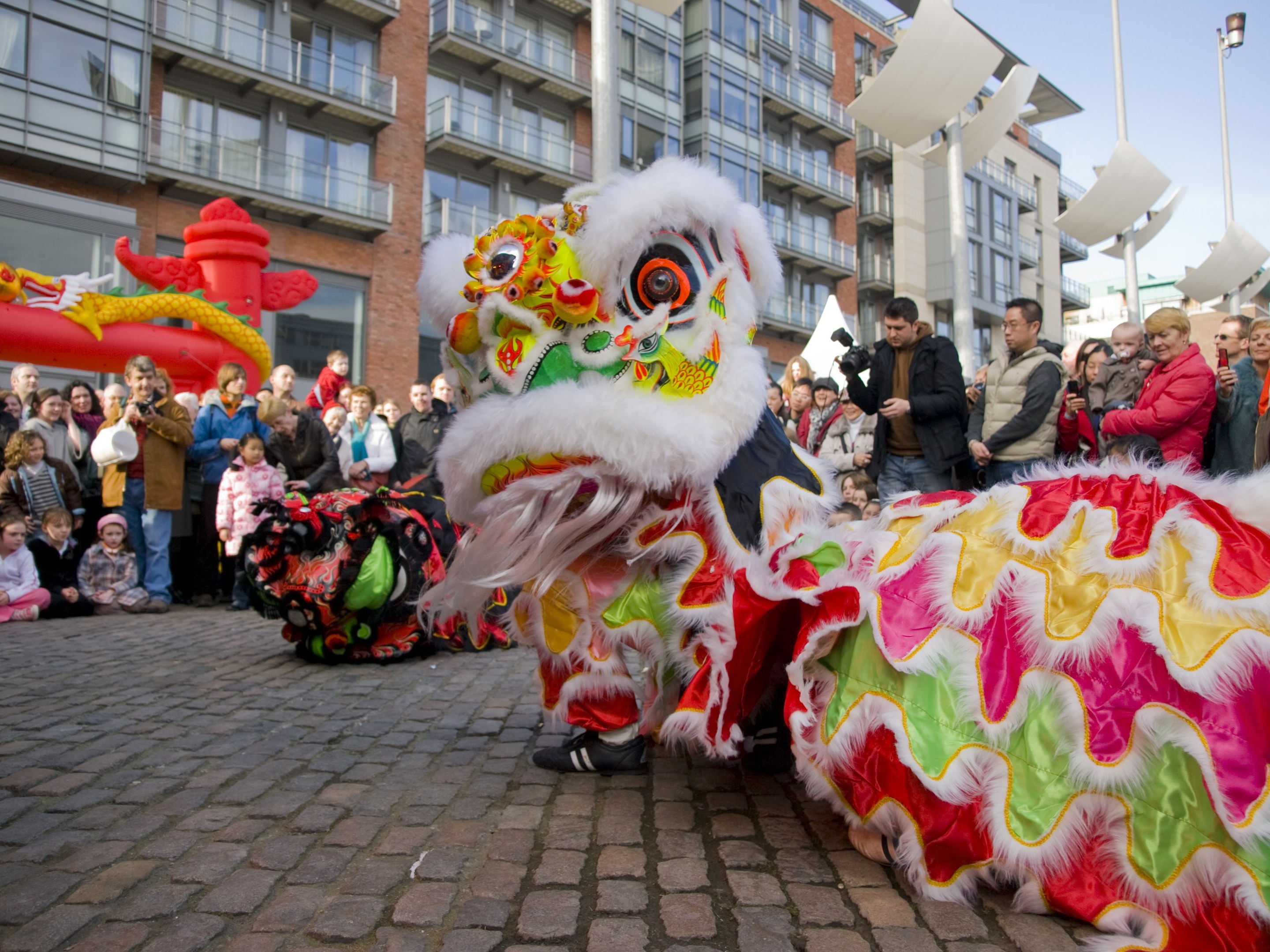 Ein lebendiges chinesisches Neujahrsfest in Amsterdam mit einem Löwen tanzen im Vordergrund und einer Menge Menschen, einige mit Kameras, drumherum, vor Buildings, Laternenmasten und einem klaren blauen Himmel.