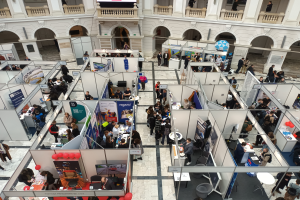 Eine große Gruppe von Menschen versammelt sich in einem Raum voller Stände, Banner, Tische, Stühle, Luftballons und einem Gebäude mit Bögen, Säulen, Geländern und einem Hintergrundbild.