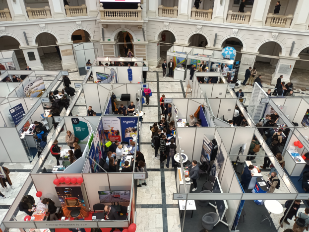 Eine große Gruppe von Menschen versammelt sich in einem Raum voller Stände, Banner, Tische, Stühle, Luftballons und einem Gebäude mit Bögen, Säulen, Geländern und einem Hintergrundbild.