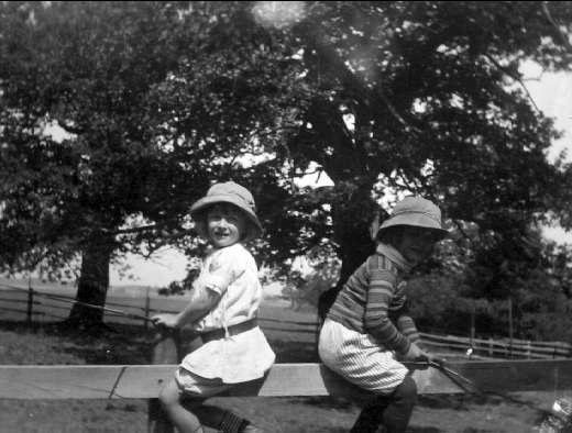 Zwei junge Jungs in Mützen halten Baseballschläger in einem grasbewachsenen Park mit Bäumen und einem Zaun im Hintergrund, die Szene in Schwarz-Weiß.