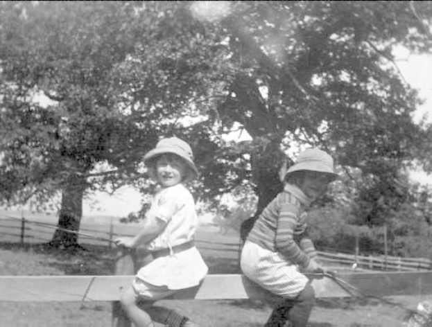 Zwei junge Jungs in Mützen halten Baseballschläger in einem grasbewachsenen Park mit Bäumen und einem Zaun im Hintergrund, die Szene in Schwarz-Weiß.