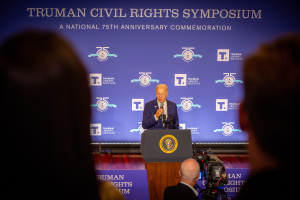 Ein Mann im Anzug und Krawatte steht an einem Rednerpult und hält ein Mikrofon in der Hand, mit einem Banner "Trumpan Bürgerrechts-Symposium" dahinter und einer Person, die eine Videokamera bedient, rechts von ihm.