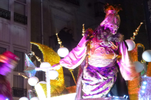 Ein Mann in einem Kostüm steht auf einem geschmückten, beleuchteten Festwagen in einer Stadtparade, mit einem Gebäude mit Fenstern und Geländern im Hintergrund.