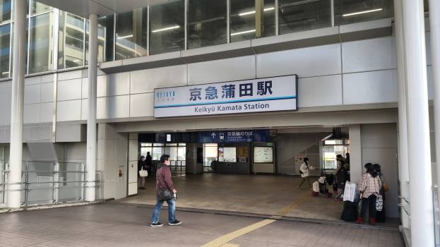 Menschen gehen durch einen Bahnhof mit überkopfschildern, Säulen, Geländern und Deckenleuchten.