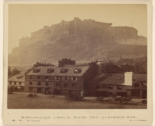Alte Fotografie des Edinburgh Castle von der Grassmarket aus betrachtet, mit Gebäuden und Karren im Vordergrund und einem Hügel im Hintergrund, mit Text unten.