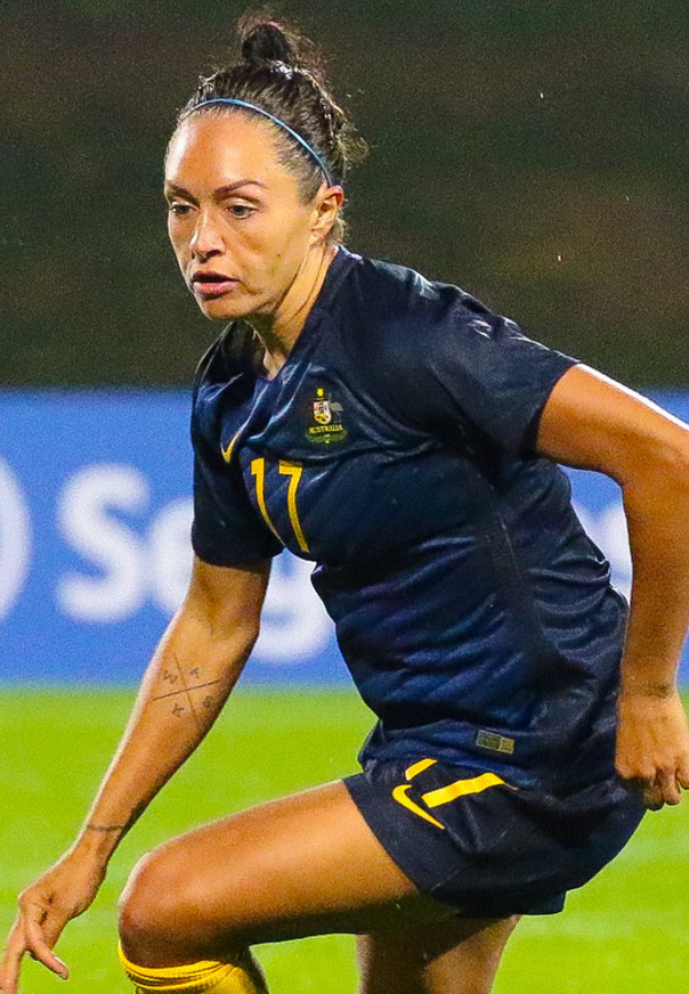Eine Fußballspielerin in einem blauen T-Shirt und gelben Shorts ist im Einsatz auf dem Feld, mit einem unscharfen Hintergrund, der eine blaue Tafel mit Text zeigt.
