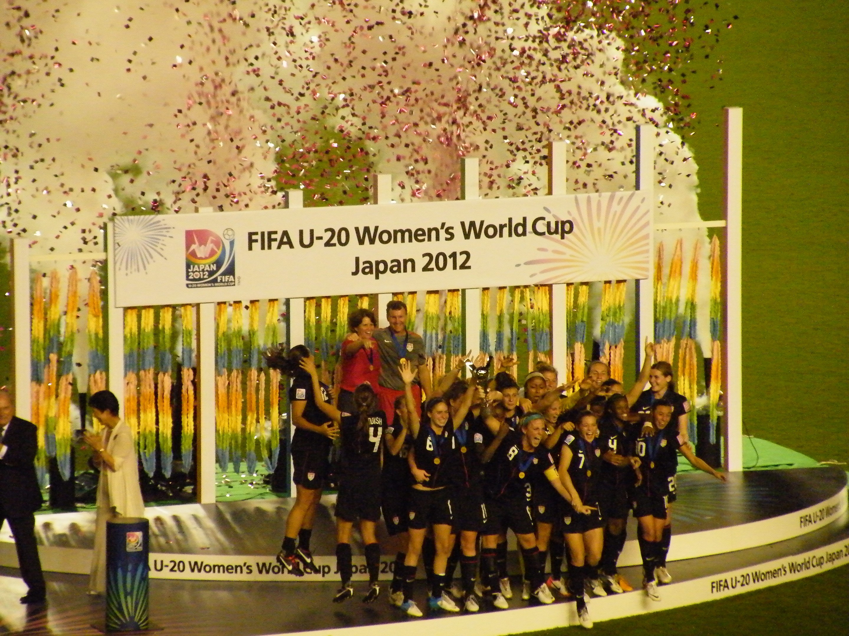 Gruppe von Frauenfußballspielerinnen in schwarzer Uniform auf einer Bühne mit Publikum und dekoriertem Hintergrund.