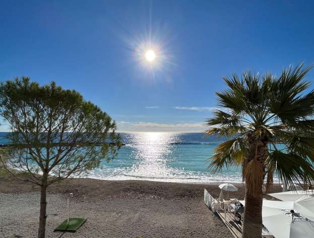 Ein Strand mit Palmen, Sonnenschirmen und üppiger Vegetation unter einem strahlend blauen Himmel mit der Sonne im Hintergrund, geeignet für eine Ferienwohnung an der französischen Riviera.